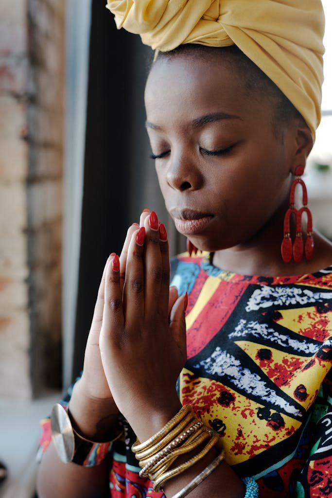 A thoughtful woman wearing colorful traditional African clothing and headwrap, praying with closed eyes.